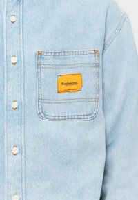 Light blue denim shirt with a button-down front, white buttons, and a chest pocket featuring a bright orange label with "Replay Inc." text.