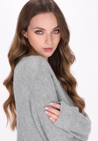 Pulover gri tricotat, cu o croială lejeră, manșete ribbate și un material moale, texturat. Modelul poartă un machiaj natural și părul ondulat.