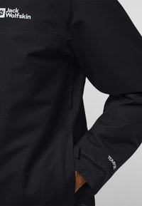 Veste imperméable noire avec une finition texturée, arborant un logo sur la poitrine, des poignets ajustables par Velcro et une coupe décontractée.