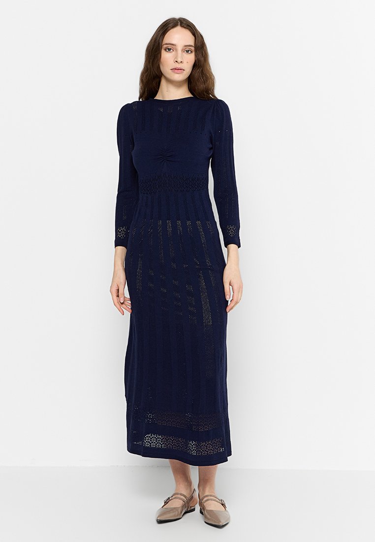 Robe en maille marine à manches longues, avec un motif texturé, des détails froncés au niveau du corsage et une silhouette droite, assortie à des ballerines marron clair.