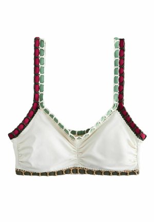 Ung kvinde med vådt hår iført en hvid crop top med grøn og lilla kant, stående udendørs med en mørk, bladfyldt baggrund.