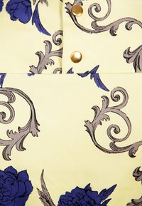 Kremowa tkanina z niebieskim kwiatowym wzorem i szarymi ornamentami, ozdobiona dwoma złotymi metalowymi guzikami ułożonymi pionowo.