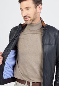 Man in een beige coltrui en een zwarte leren jas met blauwe voering, die de jas openhoudt om de binnenkant te laten zien.