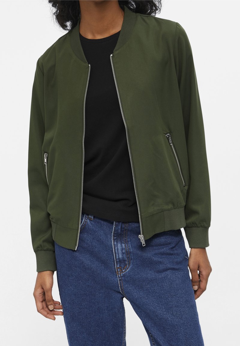 Femme portant une veste bomber vert olive avec des fermetures éclair argentées sur un haut noir et un jean bleu taille haute, debout les mains détendues.