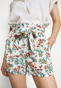 Shorts floraux avec un fond blanc, présentant un motif floral coloré vert et multicolore, une ceinture à nouer et des boutons à l'avant ; le tissu semble léger.