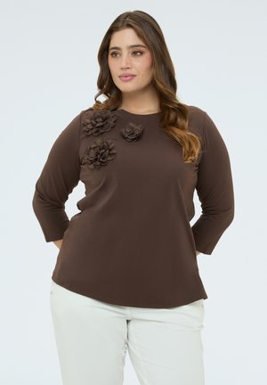 Vrouw met lang haar, gekleed in een bruine top met 3D bloemapplicaties en witte broek, poseert met handen achter de rug tegen een witte achtergrond.