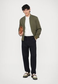 Olívazöld bomber dzseki, fehér ing, sötét nadrág, barna kézitáska és kéttónusú cipő zöld és bézs részletekkel. Egyszínű háttér.