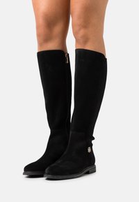Bottes noires en daim au-dessus du genou avec une semelle plate. Elles présentent une fermeture éclair latérale visible et un accent de logo doré sur le côté extérieur.