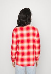 Osoba z długimi ciemnymi włosami, ubrana w czerwoną i białą koszulę w kratę, stojąca tyłem na jednolitym białym tle.