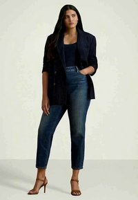 Mulher com cabelo longo e escuro, vestindo um blazer escuro riscado, top preto, jeans azuis e sandálias de salto marrons, de pé com uma mão no bolso.