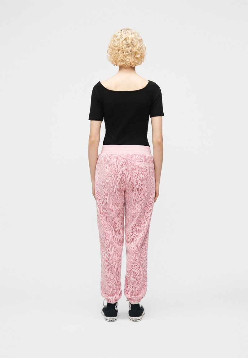 Person with short curly blonde hair stands facing away wearing a black fitted top, pink patterned pants, and black sneakers.