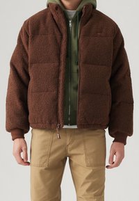 Ruskea fleece-takki, jossa on laatikkomainen muotoilu, korkea kaulus ja vetoketjusuljenta. Päällä vihreä huppari ja yhdistetty vaaleanruskeisiin housuihin.