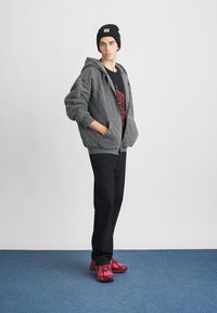 Young person wearing black beanie, gray quilted zip-up hoodie, black pants, and bright pink sneakers, standing on blue carpet against white wall.