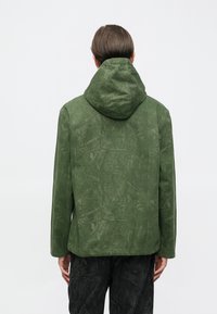 Πράσινο μπουφάν με κουκούλα και υφή, με σχέδιο εμπνευσμένο από τη φύση και χαλαρή εφαρμογή, συνοδευόμενο από σκούρο παντελόνι με μοτίβα.