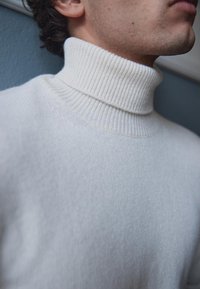 Bela turtleneck pulover iz mehkega, rebrastega pletenega materiala. Posnetek v bližini prikazuje teksturo in podrobnosti o ovratniku, z brezšivno zasnovo in ozkim prileganjem.