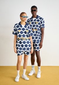 Marineblaue und weiße gemusterte Shorts und Hemden aus glattem Stoff mit einem geometrischen Design; mit gelben Logoakzenten.