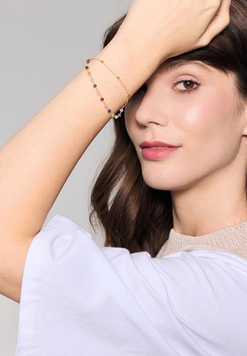Złota bransoletka z koralikami w różnych kolorach, z mieszanką okrągłych i fasetowanych kształtów, noszona na nadgarstku na neutralnym tle.