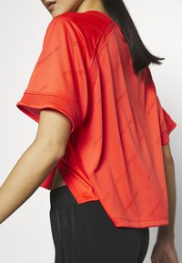 Czerwona sportowa koszulka o luźnym kroju, z krótkimi rękawami i bocznymi rozcięciami; zawiera powtarzające się logo w subtelnym wzorze.