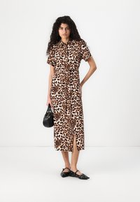 Abito maculato con maniche corte, vita cinturata e spacco frontale. Indossato con borsa nera e scarpe flat, su uno sfondo bianco semplice.