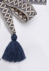 Tissu blanc avec une ceinture tissée en crème et bleu marine, ornée d'un motif géométrique et d'un détail de pompon bleu marine à l'extrémité.