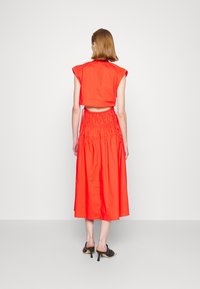 Kvinna i ärmlös röd topp och matchande högmidjad midi kjol, bärande svarta klacksandaler, stående mot en enfärgad vit bakgrund.