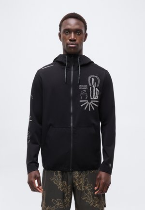 Jaunas vyras dėvi juodą užtrauktuku užsegamą džemperį su baltais grafikos dizainais ir tekstu ant krūtinės bei rankovių, derinamus su gėlių rašto šortais.