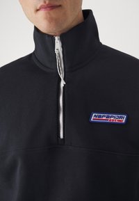 Sudadera negra con cremallera y cuello alto. Presenta una cremallera blanca, tejido texturizado y un parche de logo colorido que dice "NAPAPIJRI EST.1987."
