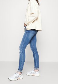Chaqueta beige claro sobre una blusa blanca, combinada con jeans skinny azules con un desgastado. Zapatillas deportivas blancas con detalles plateados completan el conjunto.