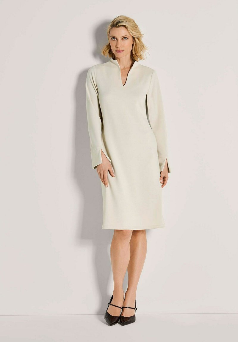 Blonde Frau in einem cremefarbenen knielangen Kleid mit langen Ärmeln und Schlitzen, die an einer hellen Wand steht, schwarze spitze Absätze.