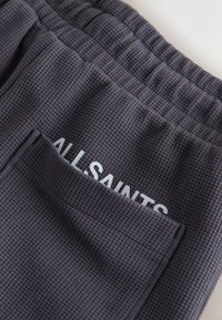 Tumedat halli tekstuuriga kangas, millel on elastne vöökoht ja tasku, kust paistab osaline valge tekst "ALLSAINTS".