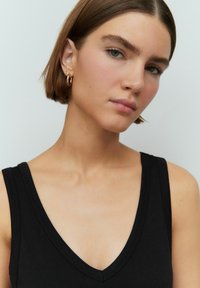 Zwarte mouwloze top met een V-hals ontwerp, gemaakt van gladde stof. Model heeft kort bruin haar en draagt gouden ringoorbellen.