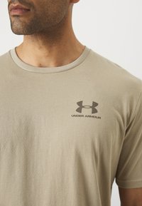 Beige bavlnené tričko s okrúhlym výstrihom, s tmavohnedým logom Under Armour na ľavej hrudi a hladkou, jemnou textúrou.
