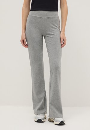 Person trägt graue weite Hosen, schwarzes Oberteil und silberne Turnschuhe und steht vor einem einfarbigen Hintergrund.