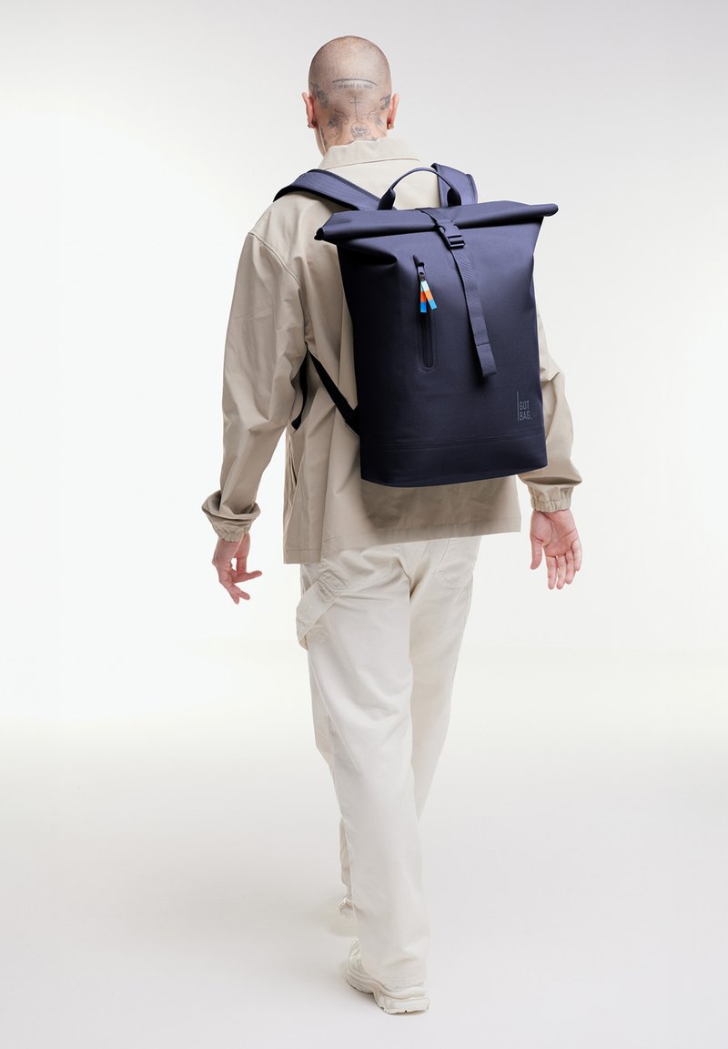 Personne en tenue beige s'éloignant, portant un grand sac à dos noir à ouverture roulante avec une poche zippée à l'avant.