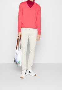 Korallfärgad v-ringad tröja över en vinröd polotröja, vita jeans, vita sneakers med orangea och marinblå accenter, håller en mönstrad tote-väska.