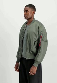 Man in olijfgroene bomberjack over een grijze shirt en zwarte broek, staande zijwaarts tegen een effen lichte achtergrond.