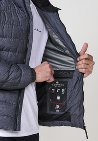 Siva puffer jakna s crnim unutrašnjim dijelom, sa mrežastom podstavom, zatvaranjem na zip, i etiketom koja detaljno opisuje upute za njegu i karakteristike.
