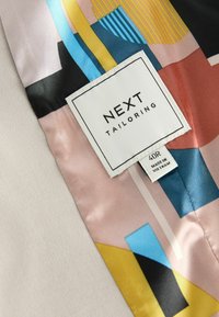 Veste sur mesure en tissu beige lisse avec une doublure en soie vibrante ornée de motifs abstraits en bleu, rose, jaune et noir. Étiquette visible.
