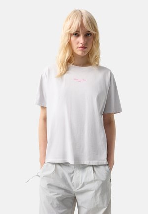 Blondynka w luźnej białej koszulce z małym różowym napisem i jasnoszarych casualowych spodniach, stojąca z rękami w kieszeniach.