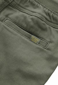 Olijfgroene broek met een gladde textuur, voorzien van een achterzak en een gele merketiket met de tekst "KENT BRAND AUTHENTIC ESTD 1982."