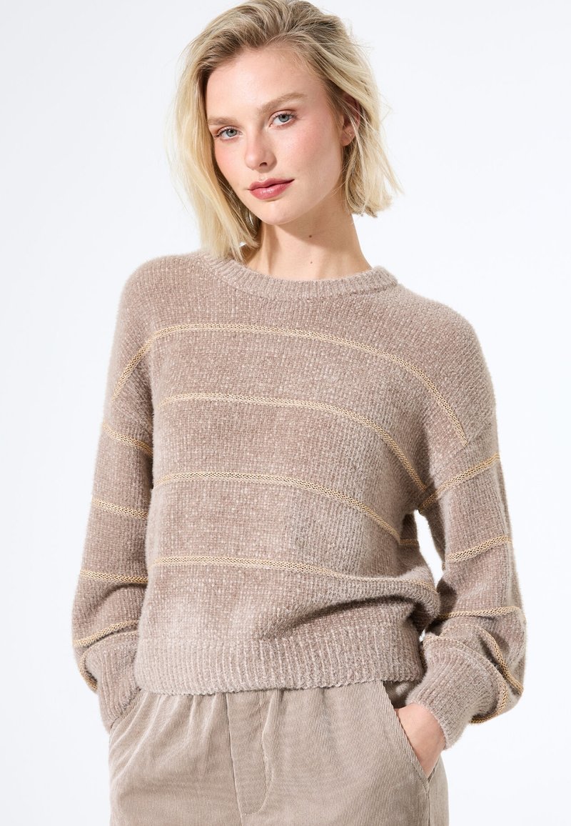 Beiger strukturierter Pullover mit goldenen Streifen, runden Ausschnitt und überschnittenen Schultern. Weiche Wolle und gerippte Saumdetails. Kombiniert mit hellen Hosen.