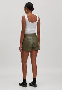 Frau in einem weißen Tanktop, olivgrünen Leder-Shorts, schwarzen Socken und schwarzen Slippern, die mit dem Rücken zu einem schlichten Hintergrund steht.