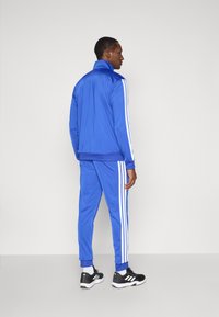 Modrý teplákový úbor zo hladkého materiálu, so bielymi pásmi po bokoch bundy aj nohavíc. Obsahuje vysoký golier a žebrované manžety.