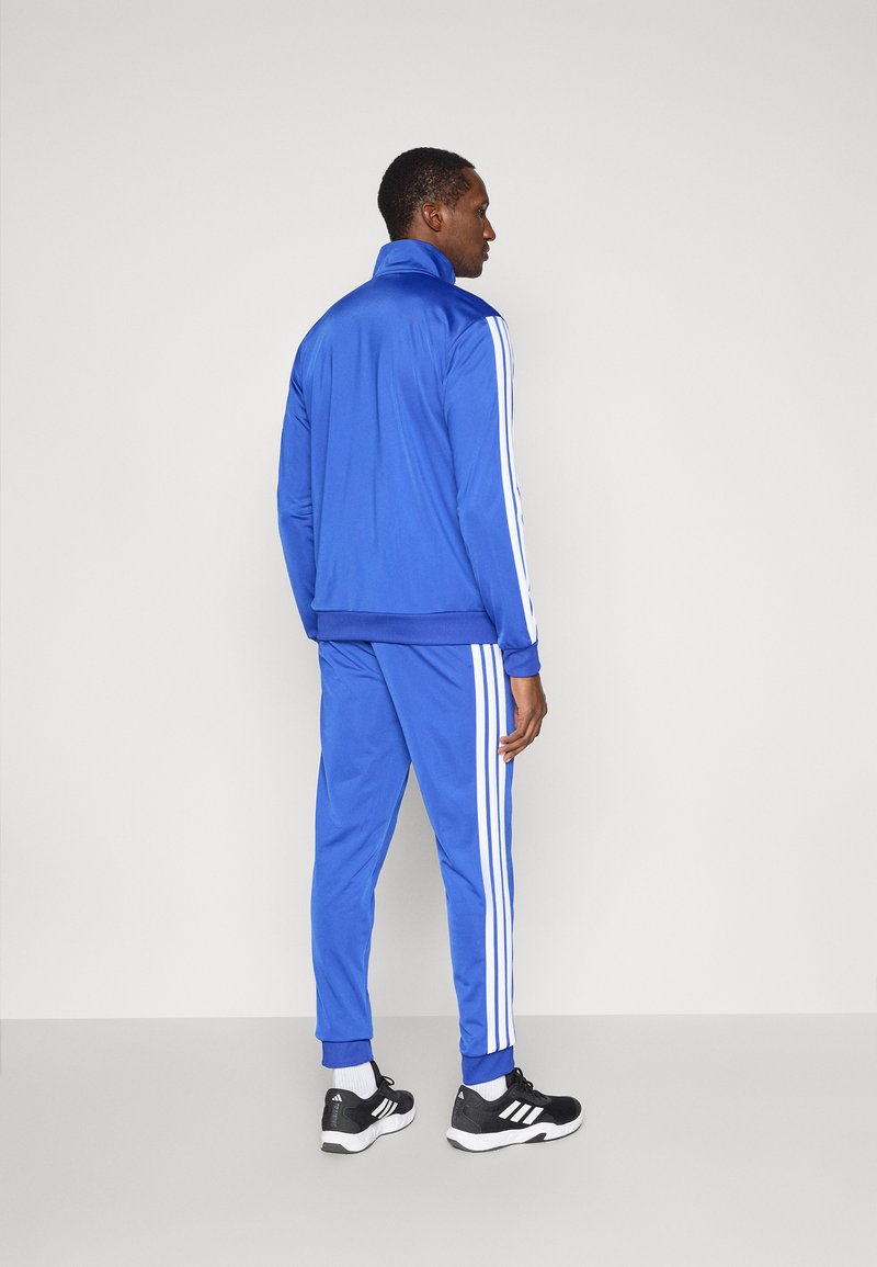 Blauer Trainingsanzug aus glattem Stoff, mit weißen Streifen an den Seiten sowohl der Jacke als auch der Hose. Beinhaltet einen hohen Kragen und gerippte Bündchen.