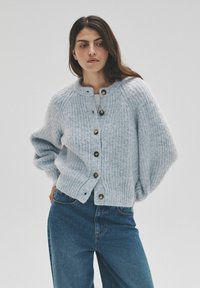 Gaiši zila adīta kardigana ar apaļu izgriezumu, ribota tekstūra un lielām brūnām pogām, valkāta kopā ar zilām džinsu biksēm.