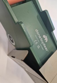 Contenedor de plástico verde con un acabado suave, con el logo "donebydeer", parcialmente visible dentro de una caja de cartón marrón.