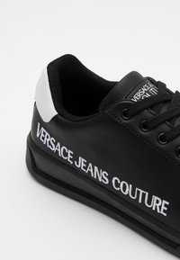 Svart sneakers i konstläder med vit logotypdetalj, texturerad tygfoder och platt svart gummisula; snörningsdesign.