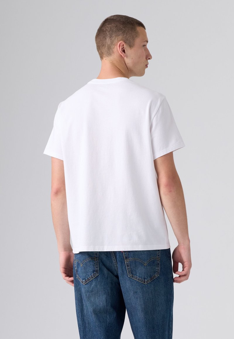 Young man with short hair wearing a plain white t-shirt and blue jeans, standing facing away against a light gray background.