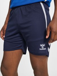 Prikaz u krupnom planu tamnoplave atletske kratke hlače s bijelim chevron prugama i hummel logotipom na bedru koje nosi osoba s rukama opuštenim uz bokove.