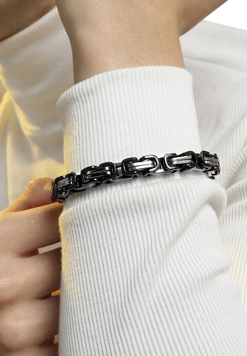Silberne und schwarze Metallkette Armband mit miteinander verbundenen Gliedern, die auf einem weißen gerippten Pulloverärmel liegt. Glatte Oberflächenstruktur, elegantes Design.